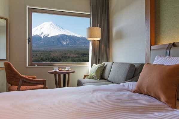 A hotel room with a bed, a sofa, a chair, a small table with a cup and saucer, and a view of a snow-capped mountain through a large window. - Highland Resort Hotel & Spa (Fujiyoshida)