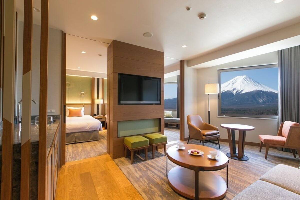A hotel room with a bed, a TV, a seating area with a table, and a large window with a mountain view.