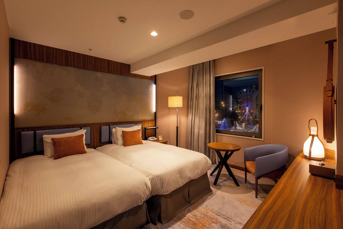 A hotel room with two beds, a desk, a chair, a lamp, and a window with a night view.
