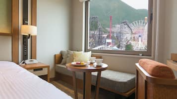 A hotel room with a bed, a bedside table with a lamp, a small table with a cup and plate, and a view of a ferris wheel and mountains through the window.
