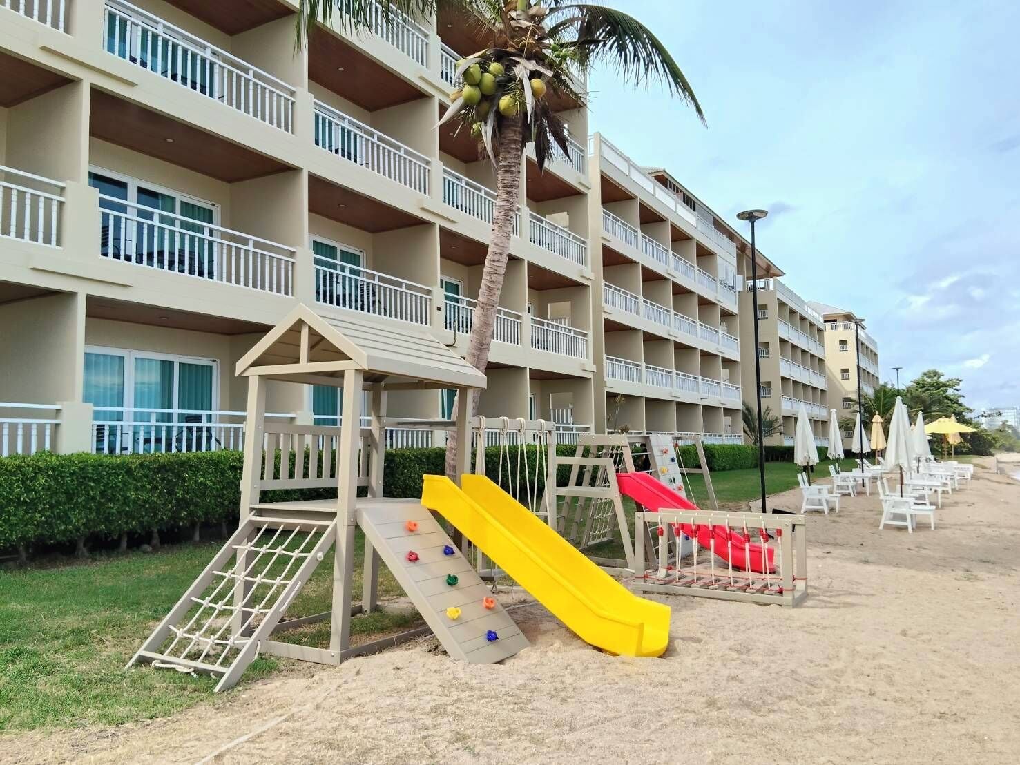 A multi-story hotel with balconies, a playground with slides, and a palm tree.