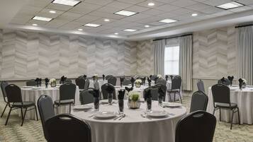 Un salón de conferencias con varias mesas redondas, cada una con platos, cubiertos y jarrones de flores. La habitación tiene un papel tapiz y ventanas grandes con cortinas.