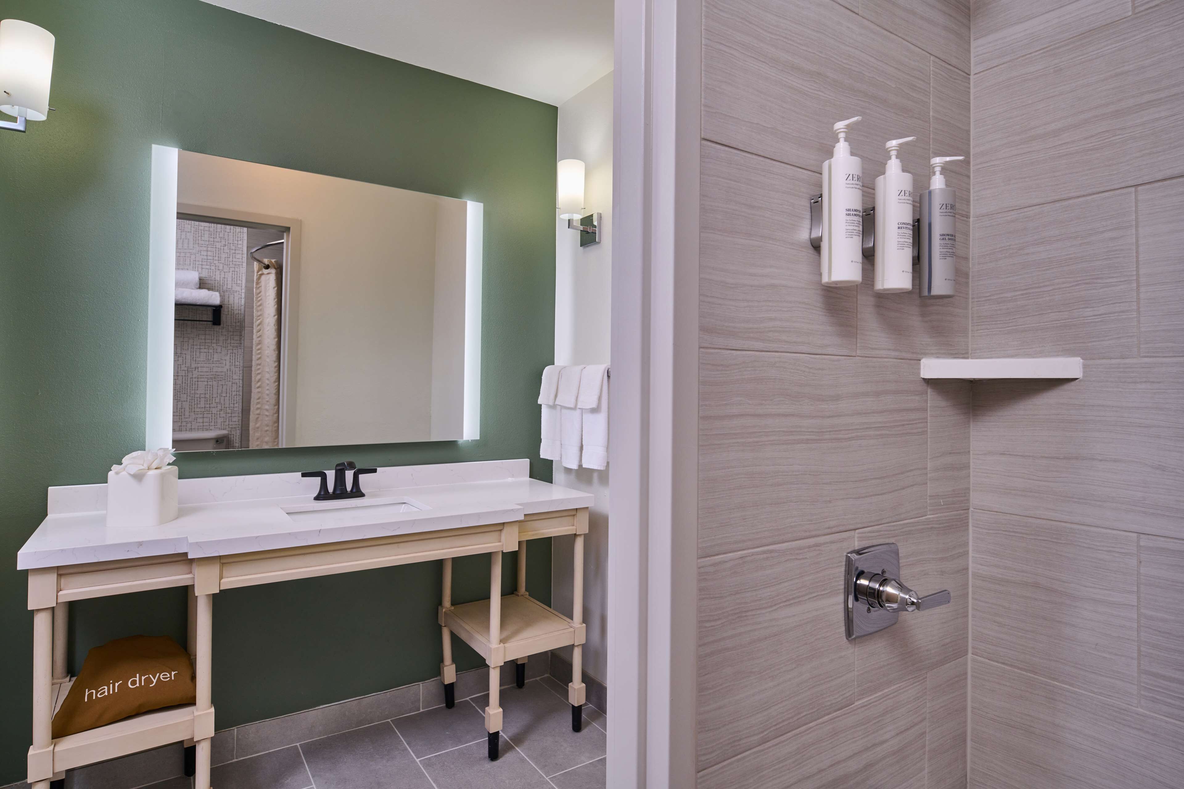 Une salle de bain avec un meuble-lavabo blanc, un sèche-cheveux et un espace douche équipé de produits de toilette.