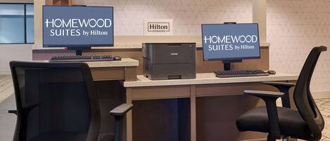 Un comptoir d’accueil avec deux écrans affichant la mention « HOMEWOOD SUITES by Hilton » et un panneau « Hilton HONORS ». On y trouve une imprimante Brother et deux chaises de bureau noires.