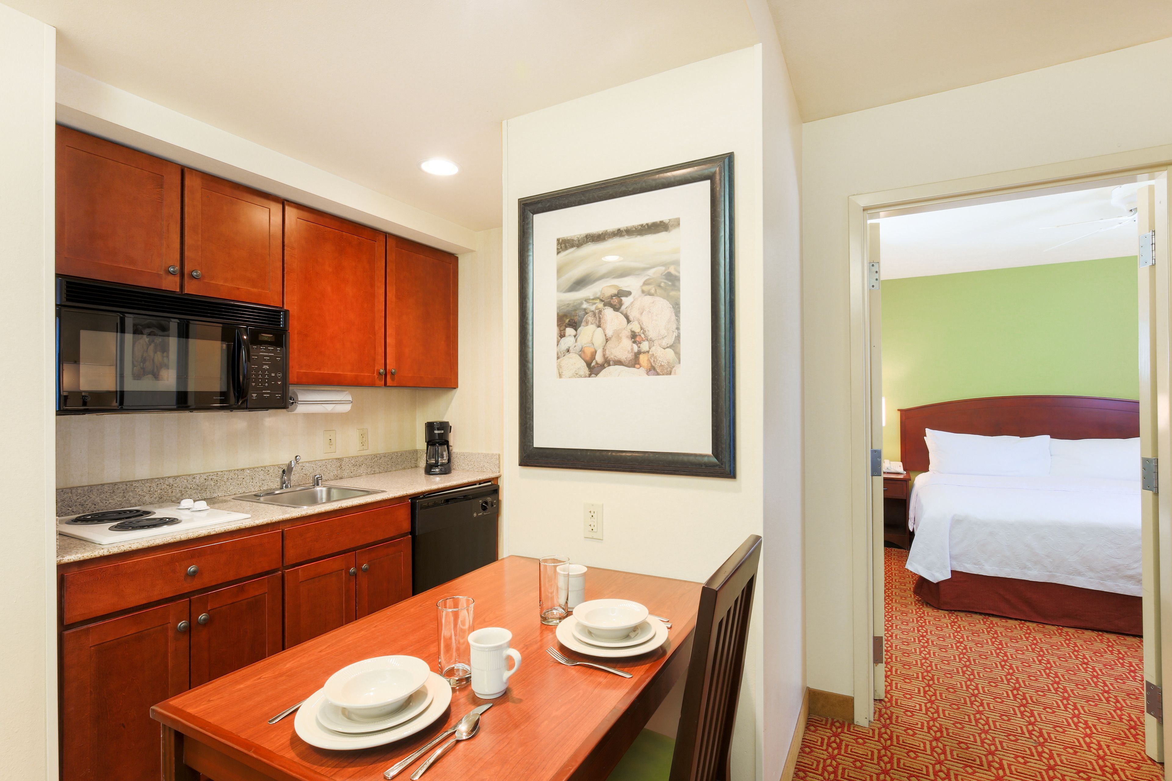 A hotel room with a kitchenette, a bed, and a framed picture on the wall.