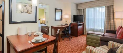 A hotel room with a dining table set for two, a television, a sofa, and a patterned carpet.