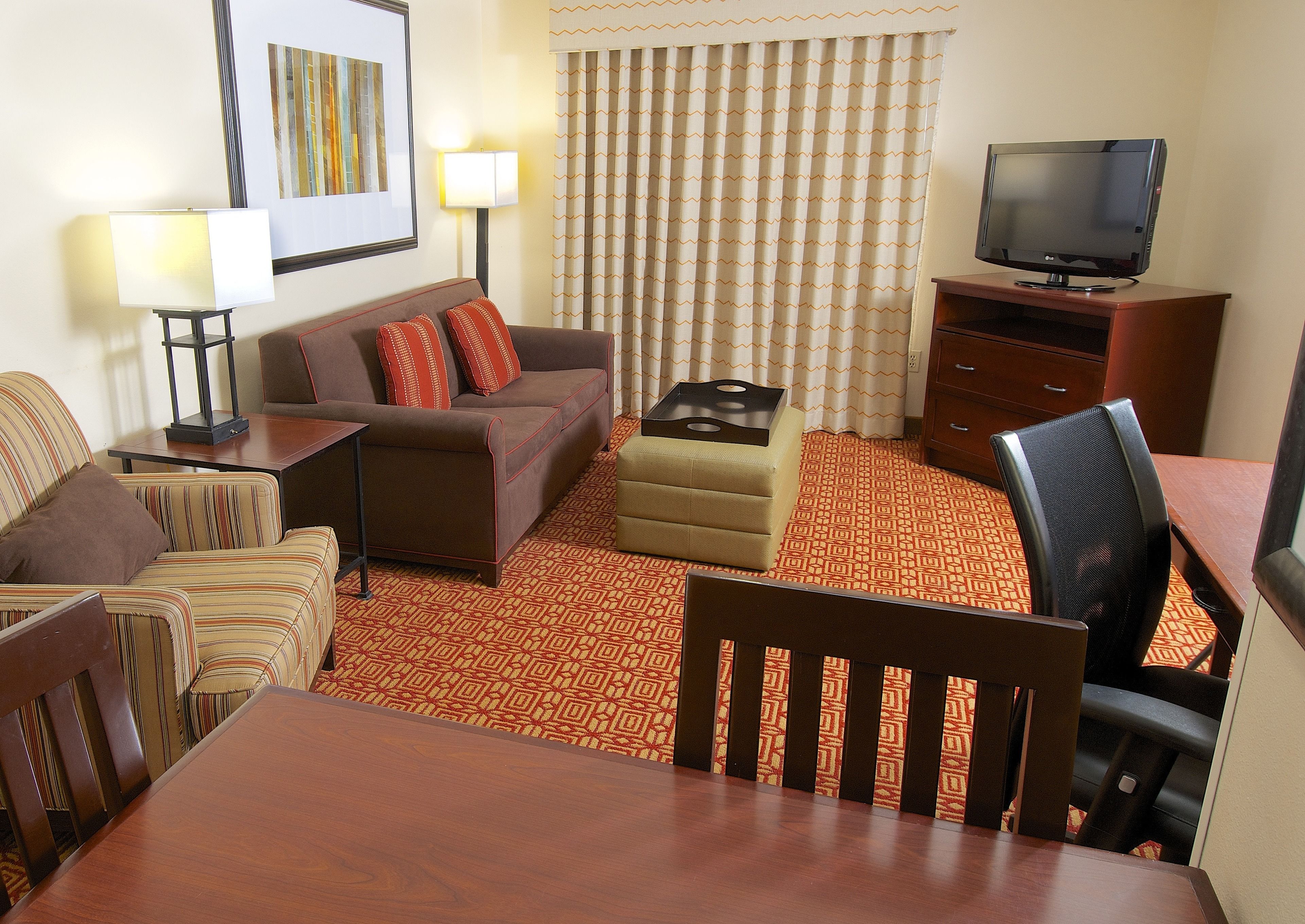 A hotel room with a sofa, armchairs, a dining table, a TV, and a patterned carpet.