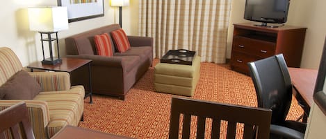 A hotel room with a sofa, armchairs, a dining table, a TV, and a patterned carpet.