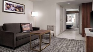 A modern hotel room with a gray sofa, a small armchair, a wooden coffee table, a framed artwork on the wall, a lamp, and a view of the city through the window. - Hilton Garden Inn Detroit Downtown (Detroit)