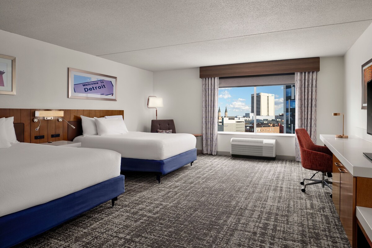 A hotel room with two beds, a desk, a chair, a window with a city view, and a sign reading "Welcome to Detroit".