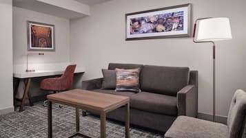 Ein modernes Hotelzimmer mit einem grauen Sofa, einem Holztisch, einem roten Sessel, einem Schreibtisch und einer Stehlampe.