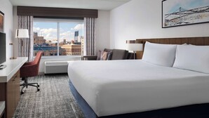 A hotel room with a large bed, a desk with a chair, a window with a view of buildings, and a painting on the wall. - Hilton Garden Inn Detroit Downtown (Detroit)