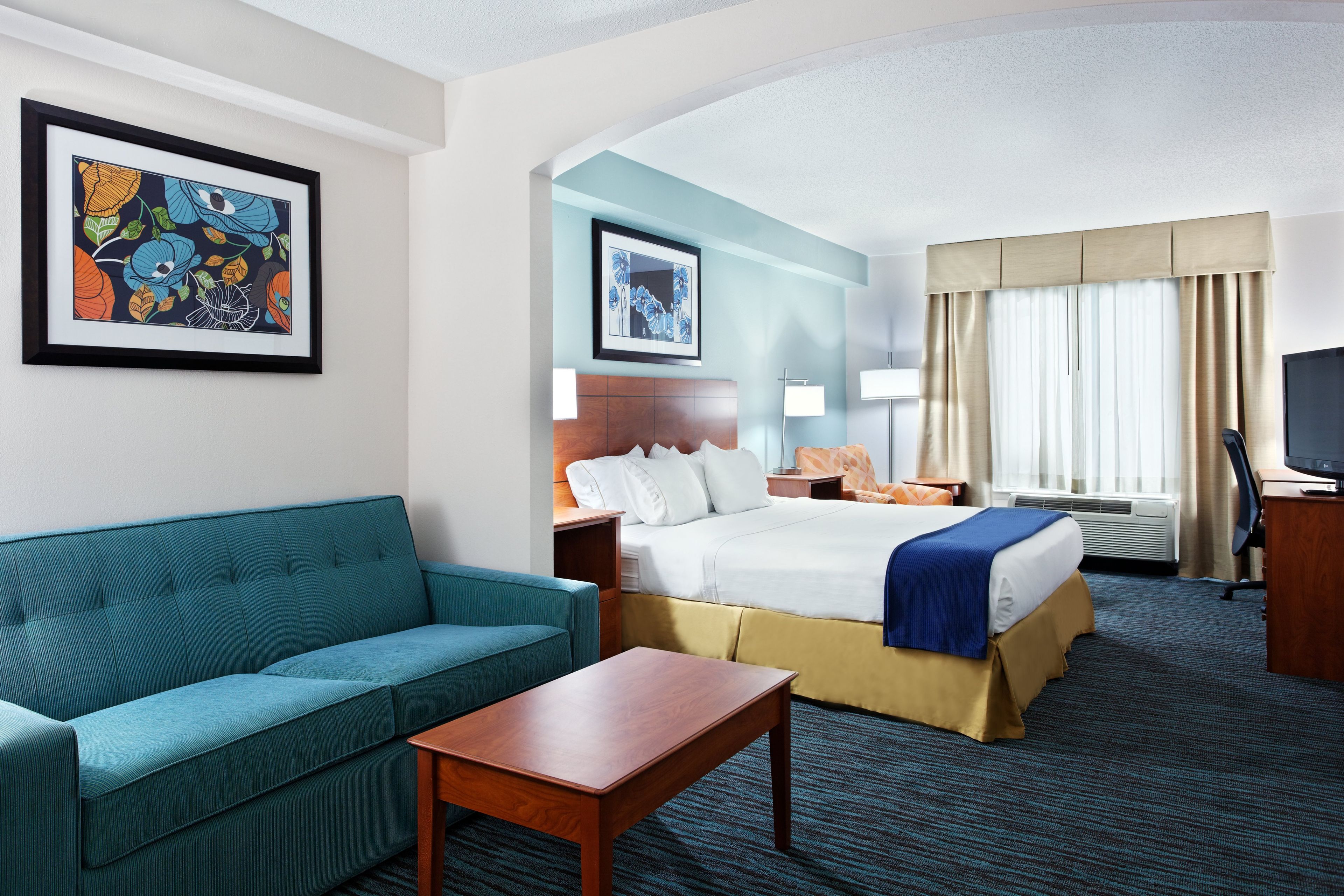 Ein Hotelzimmer mit einem Bett, einem Sofa, einem Schreibtisch, einem Fernseher und zwei gerahmten Kunstwerken an der Wand.