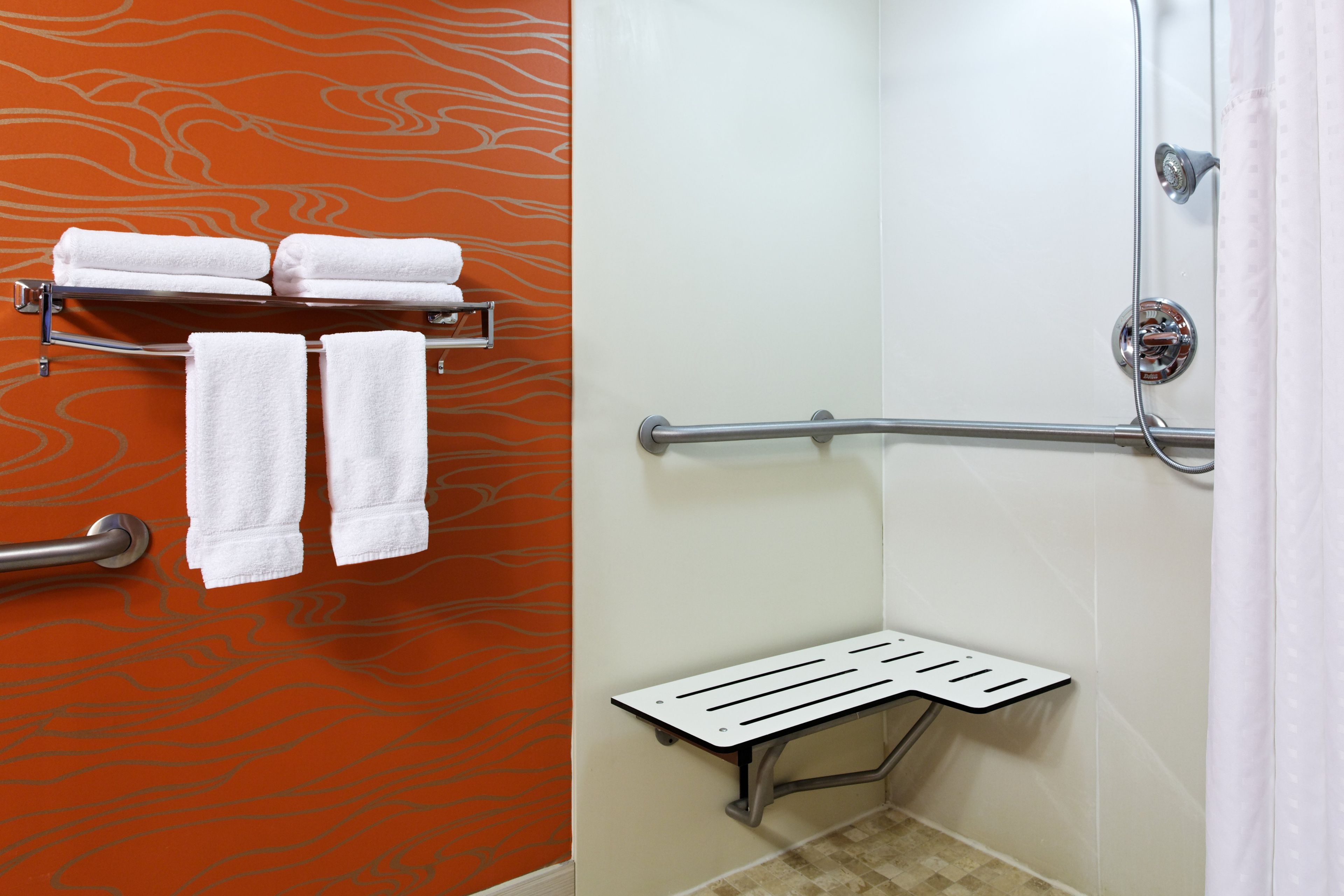 A bathroom with a shower, a towel rack with towels, a grab bar, and a shower seat.