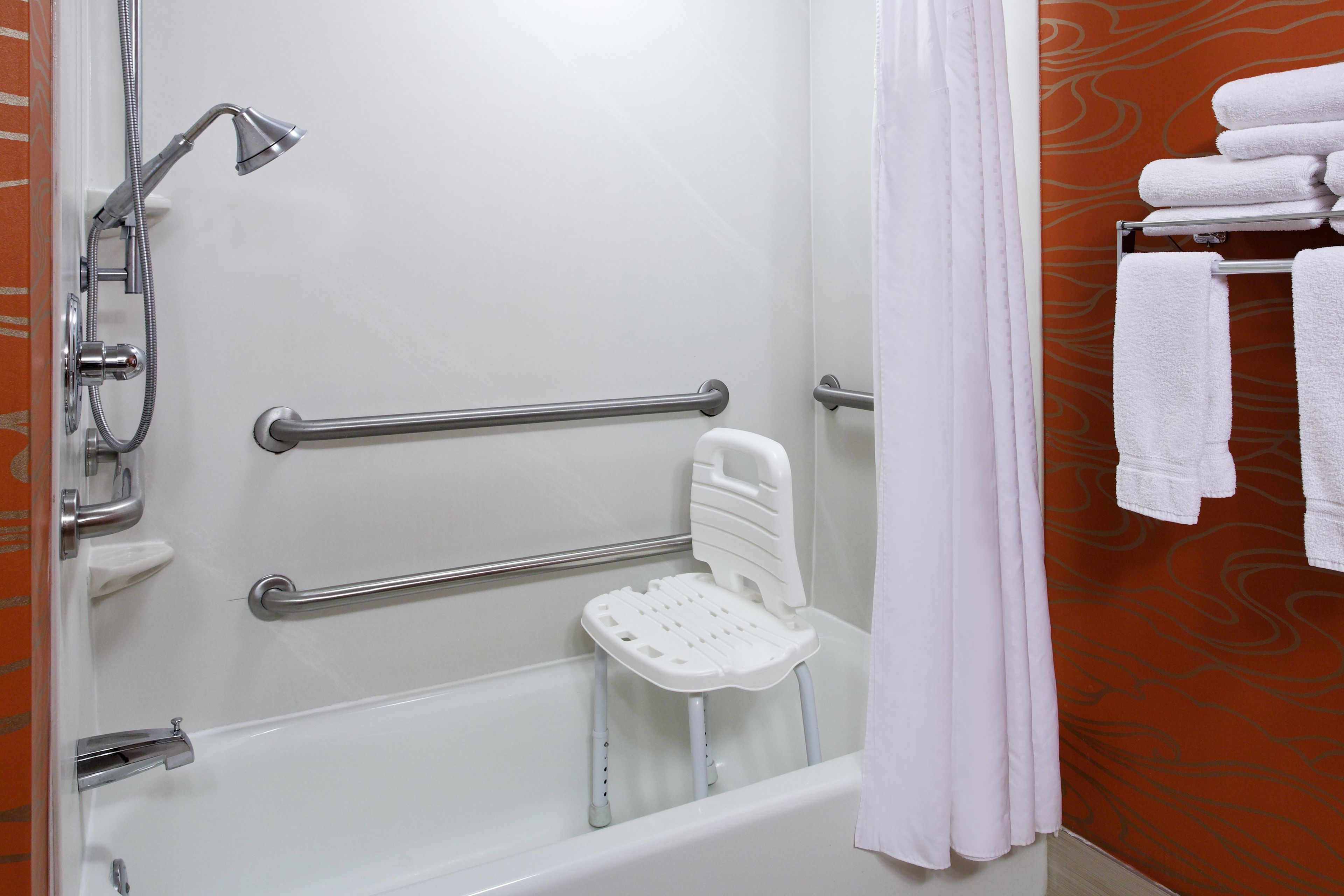 Ein Badezimmer mit einer weißen Badewanne, Haltegriffen, einem weißen Duschstuhl und einem Handtuchhalter mit gefalteten Handtüchern.