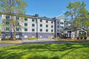 A multi-story hotel with a central entrance, parking lot, and surrounding greenery.