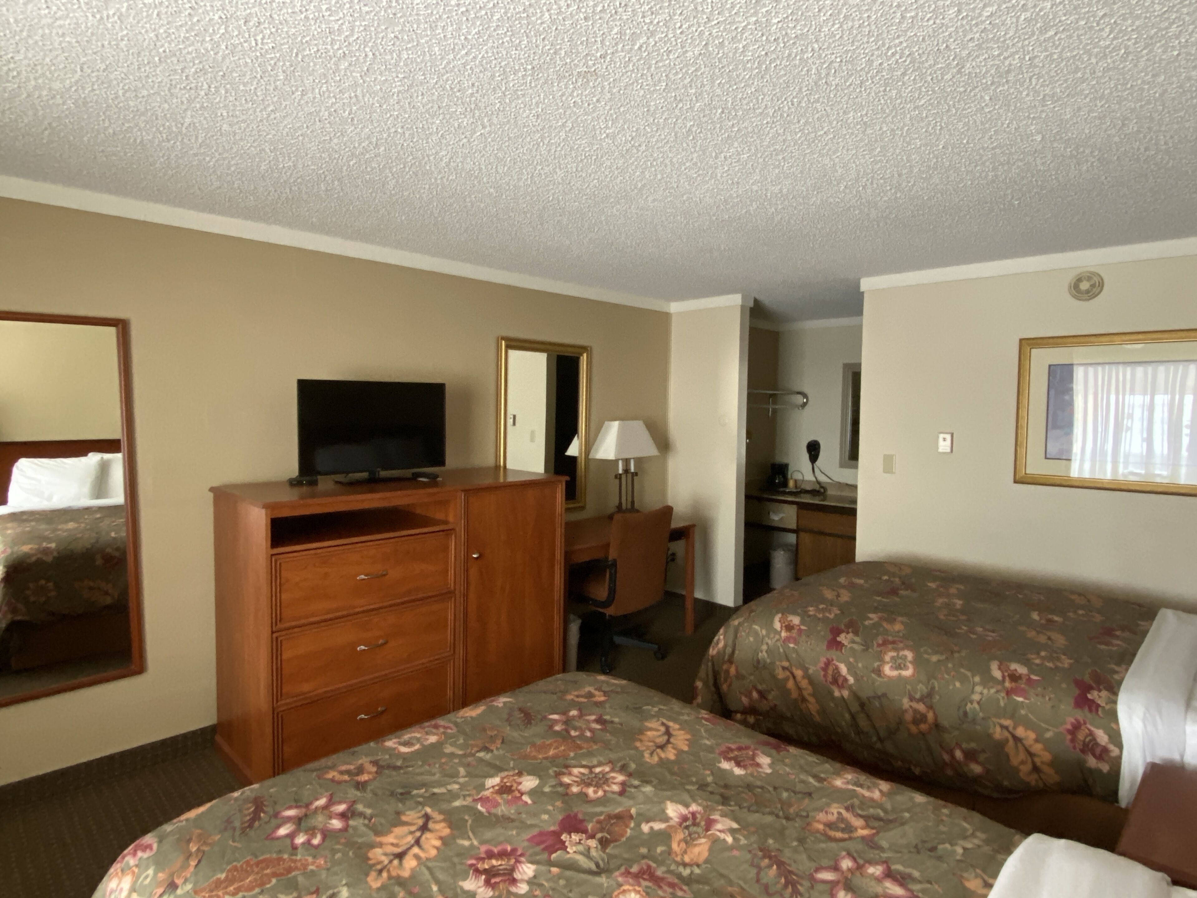 A hotel room with two beds, a TV on a wooden dresser, a desk with a lamp, and a mirror.