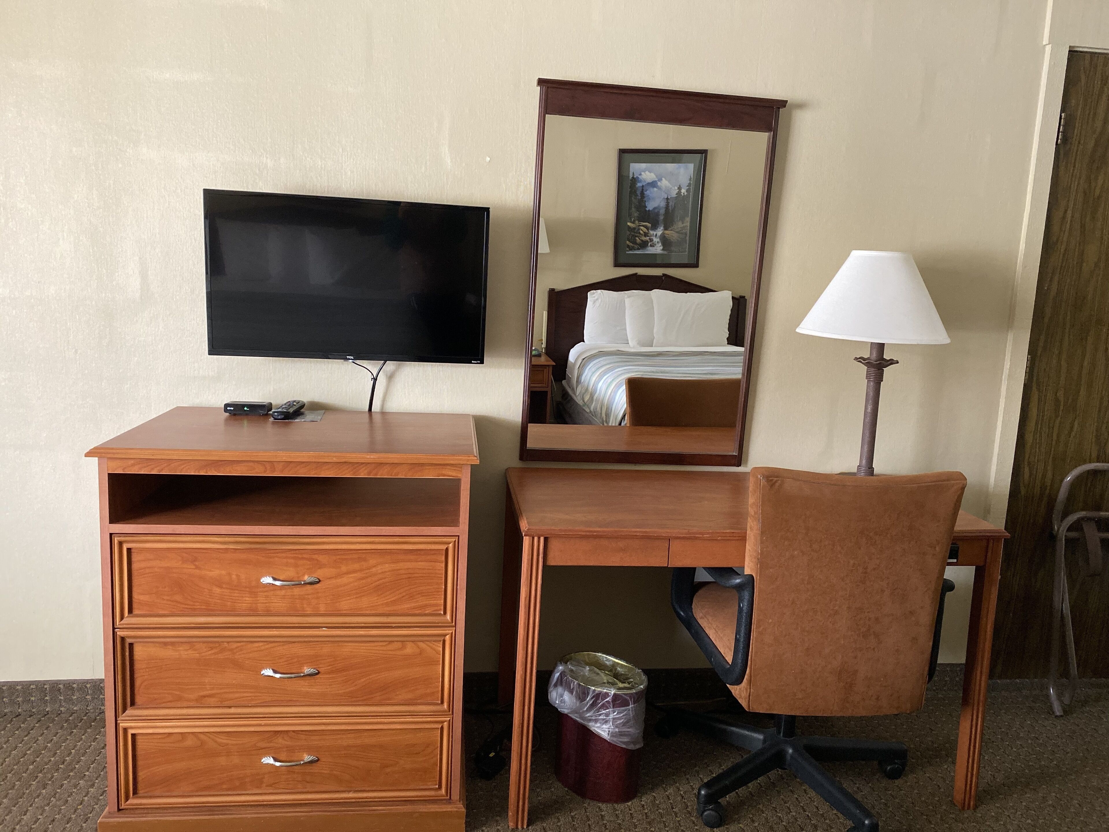 Une chambre d’hôtel avec un bureau en bois, une télévision, un lit avec une tête de lit, une lampe et une poubelle.