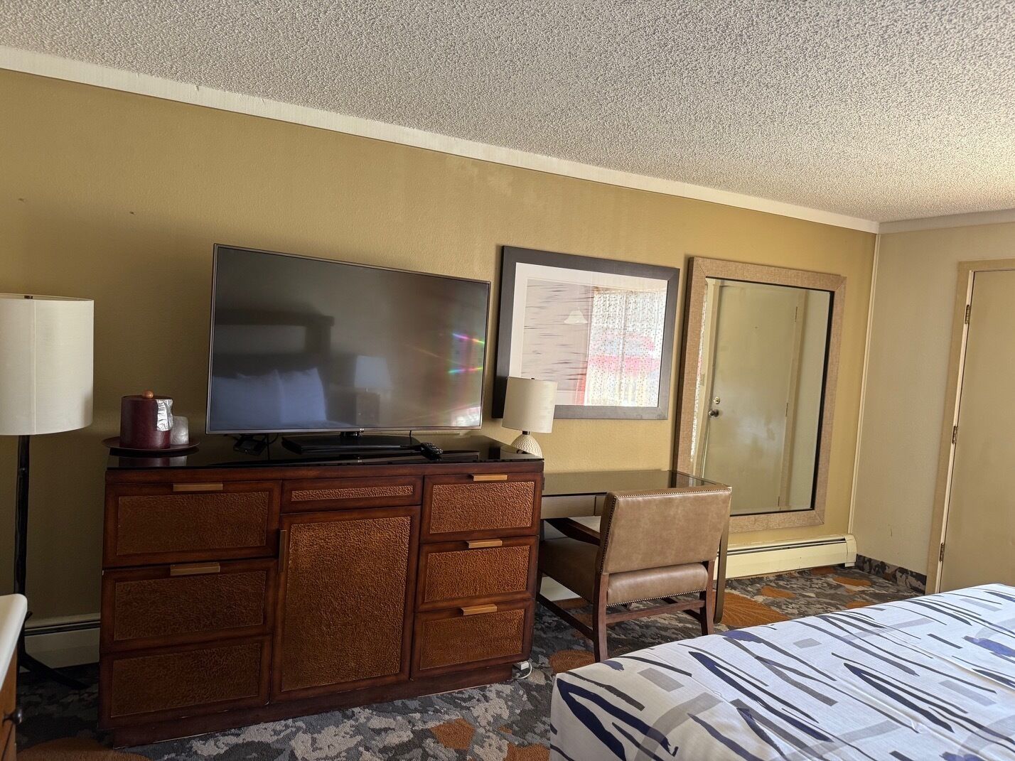 A hotel room with a flat-screen TV on a wooden dresser, a chair, a bed with a patterned bedspread, a lamp, and a door with a mirror.