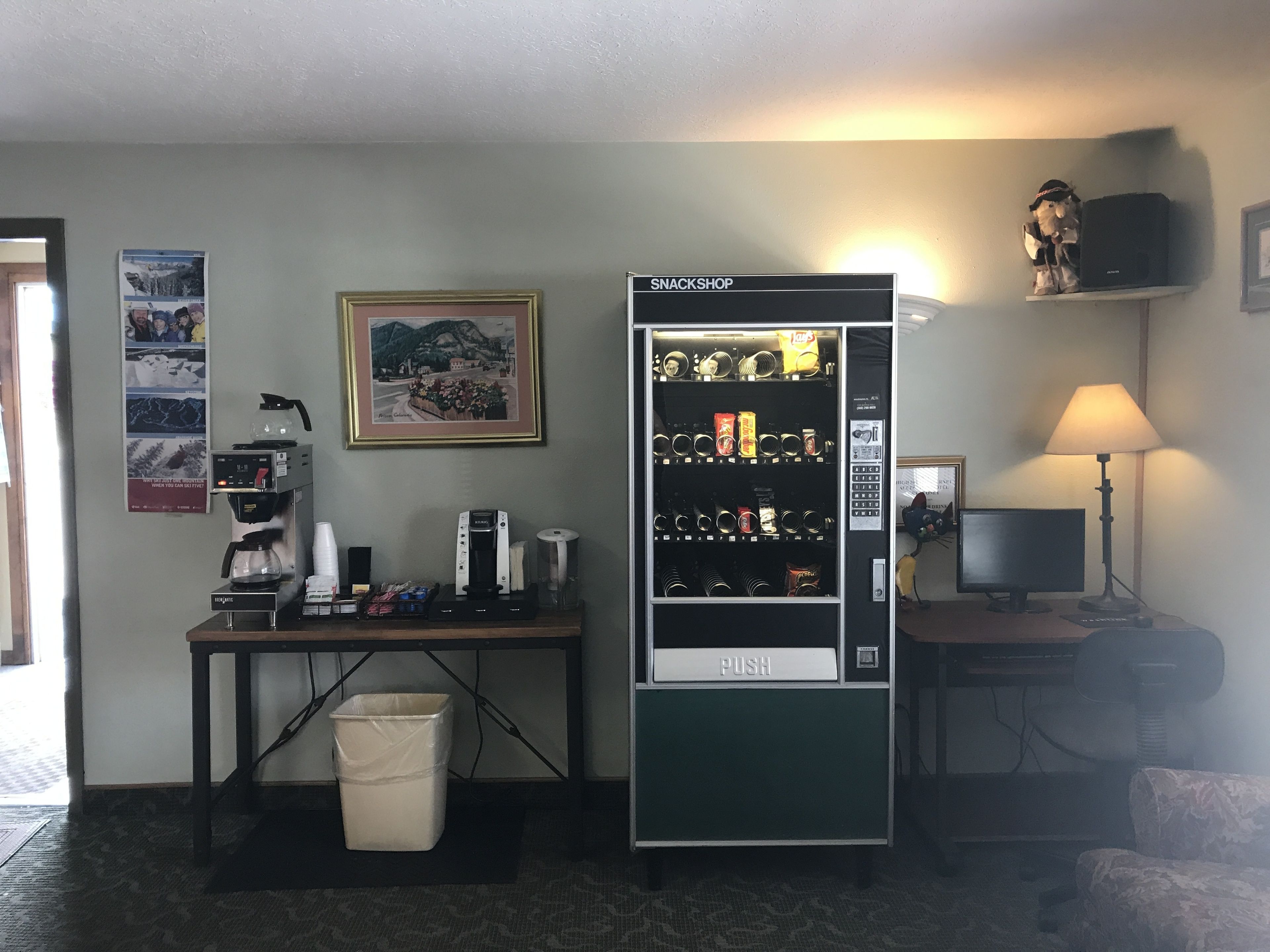 Una habitación con cafetera, máquina expendedora, escritorio, ordenador y lámpara.
