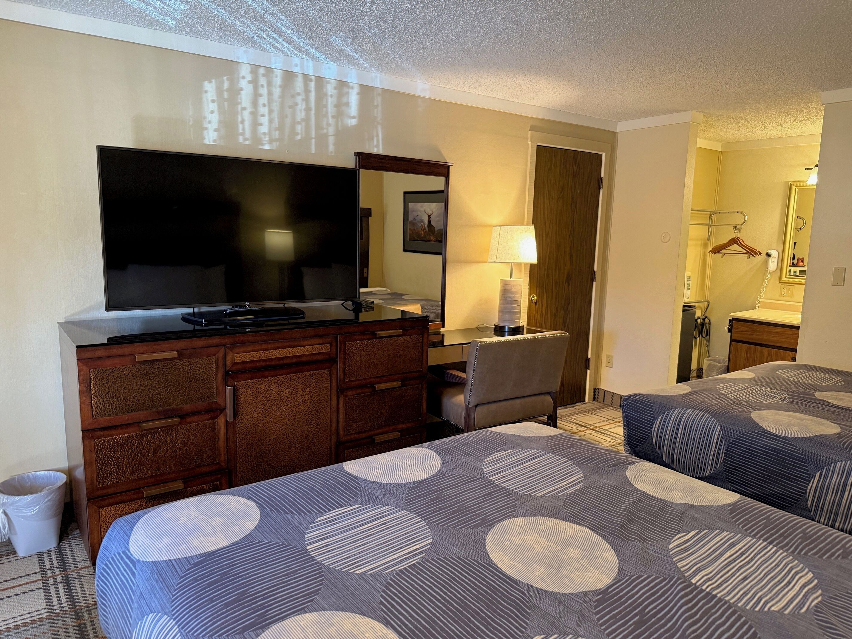 A hotel room with a large flat-screen TV, a desk with a lamp, two beds with patterned bedding, and a bathroom visible through an open door.