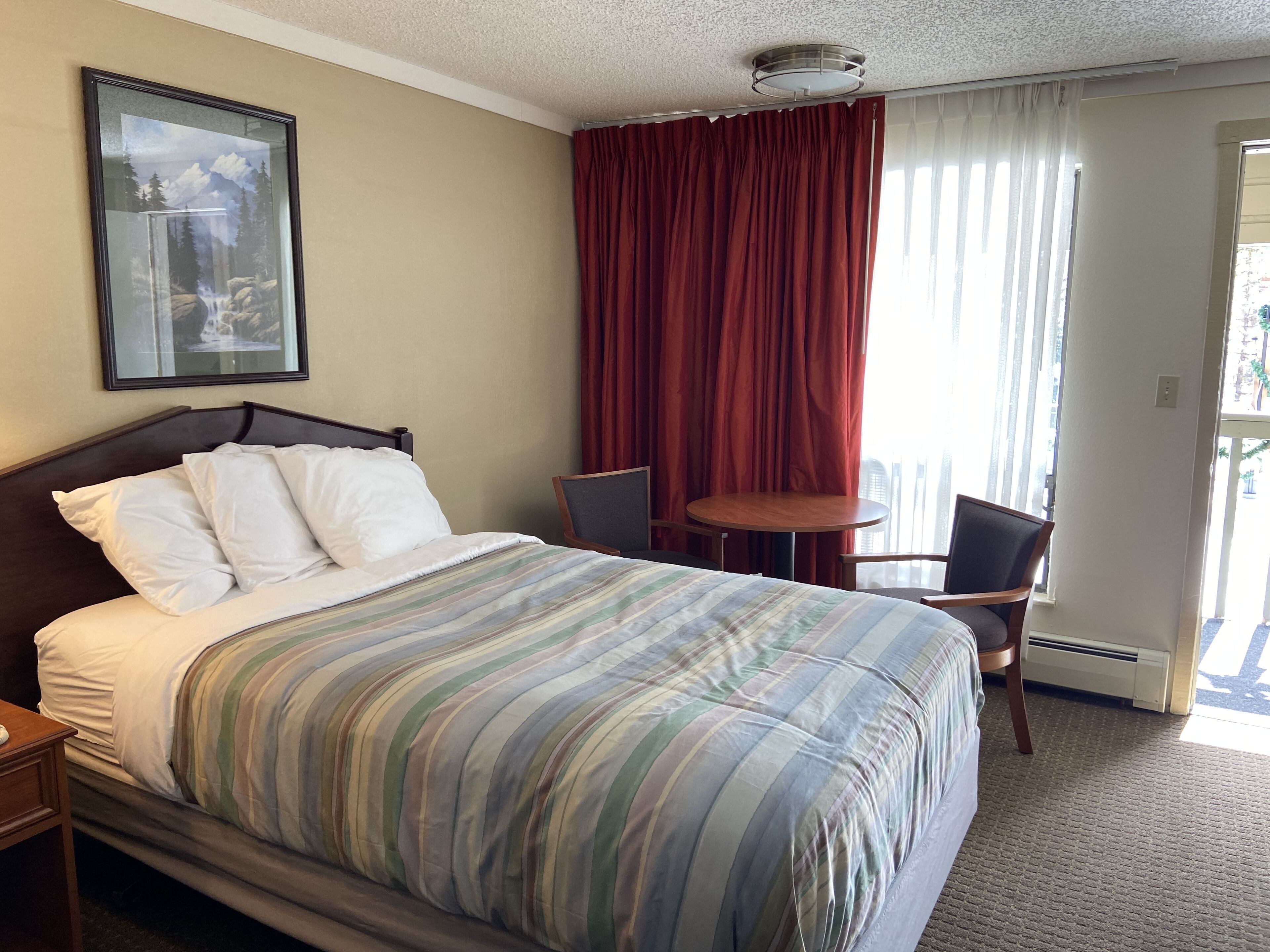 Habitación de hotel con cama, una mesita, dos sillas, ventana con cortinas y un cuadro en la pared.