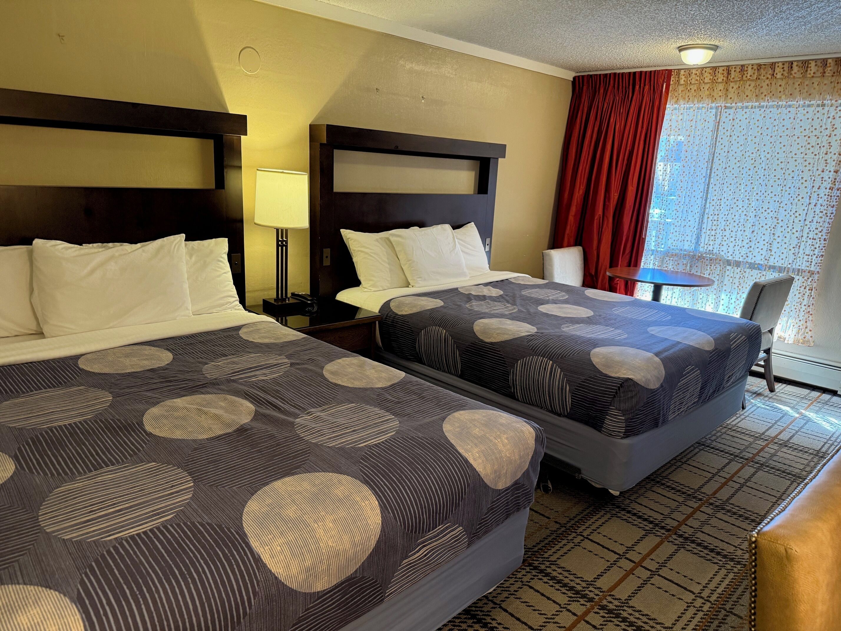 A hotel room with two beds, a desk, a chair, a lamp, and a window with curtains.