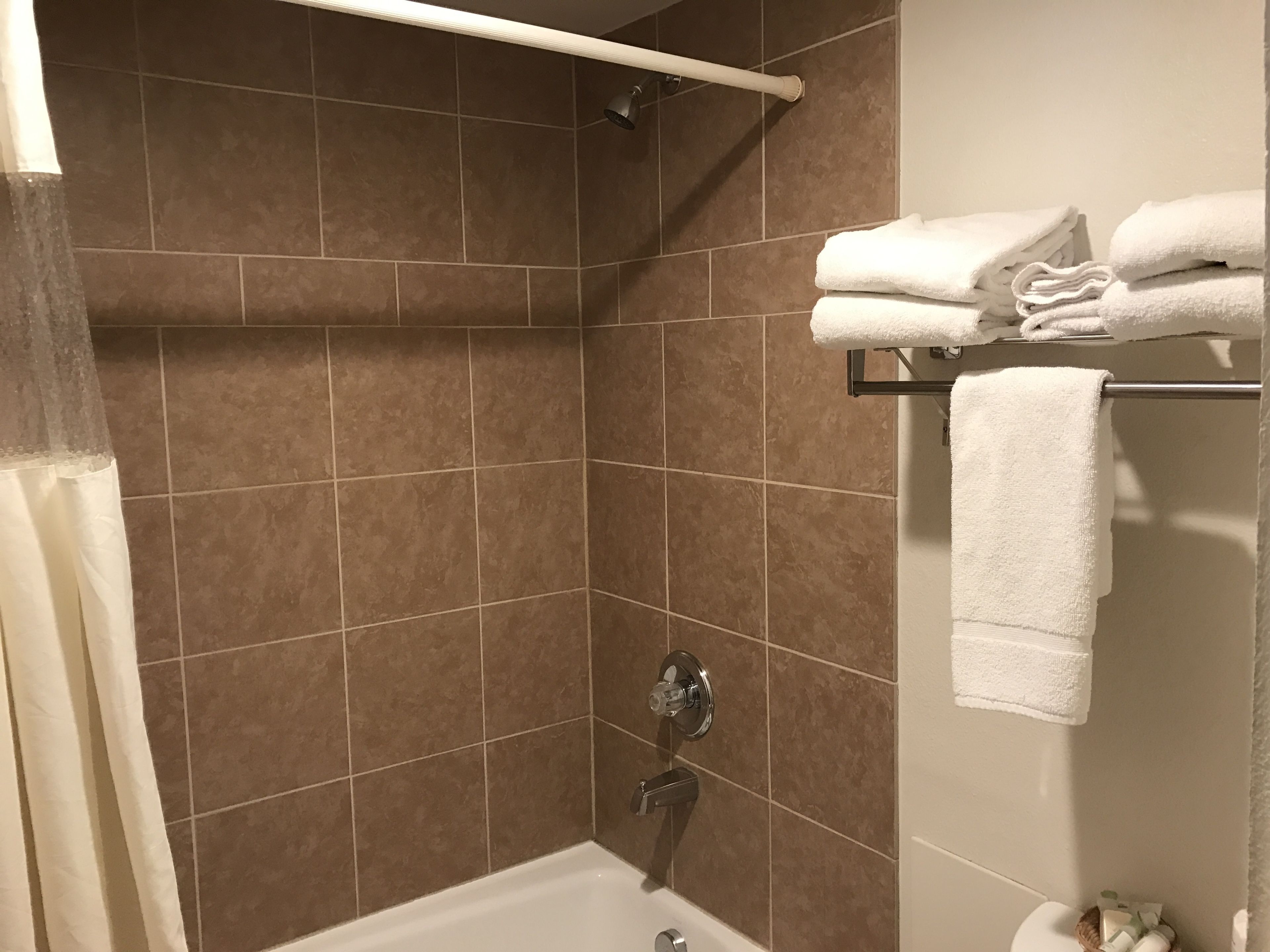 A bathroom with brown tiles, a white bathtub, a towel rack with folded towels, and a showerhead.