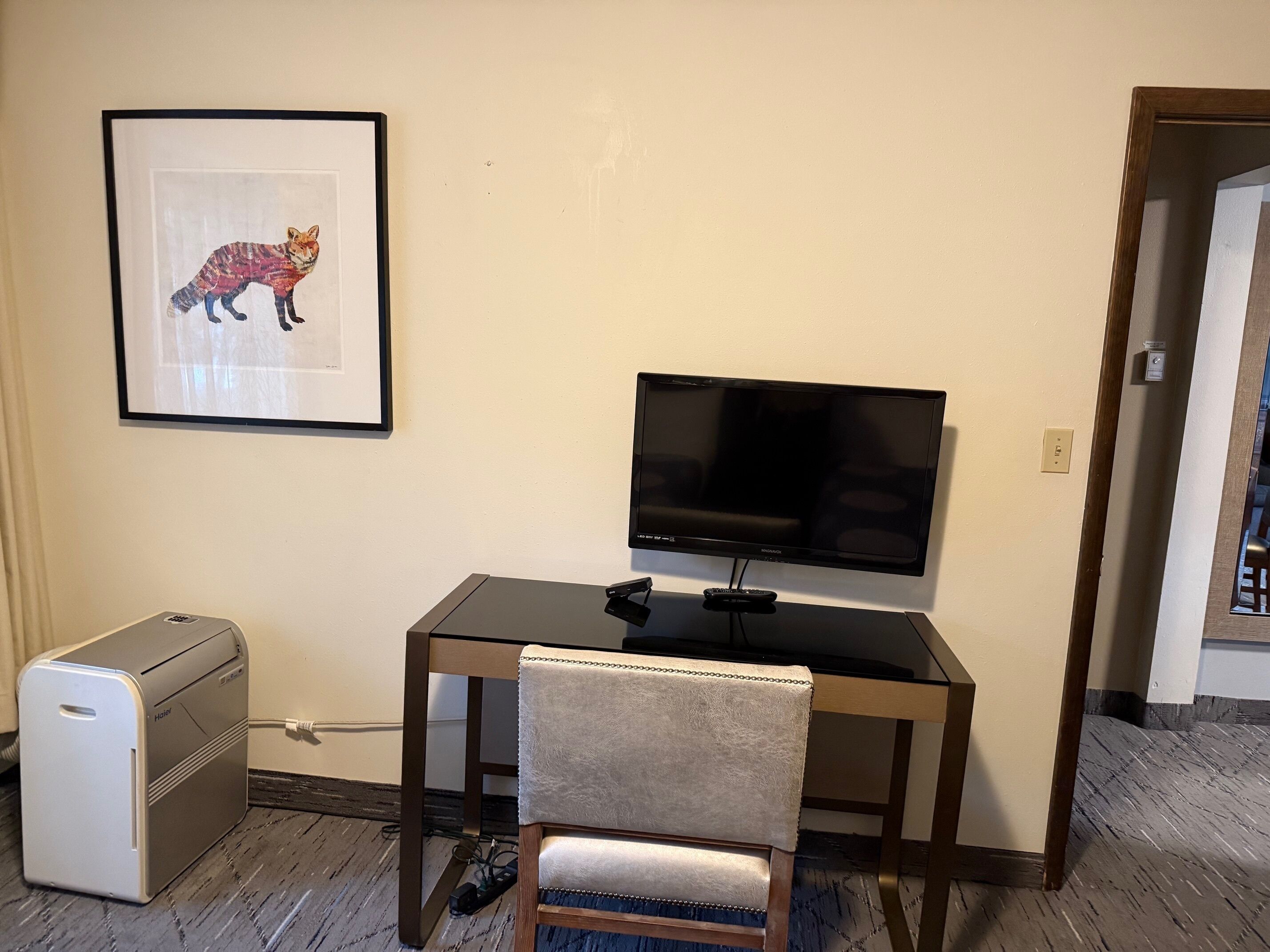 A hotel room with a desk, a flat-screen TV, a framed picture of a fox, a mini-fridge, and a chair.