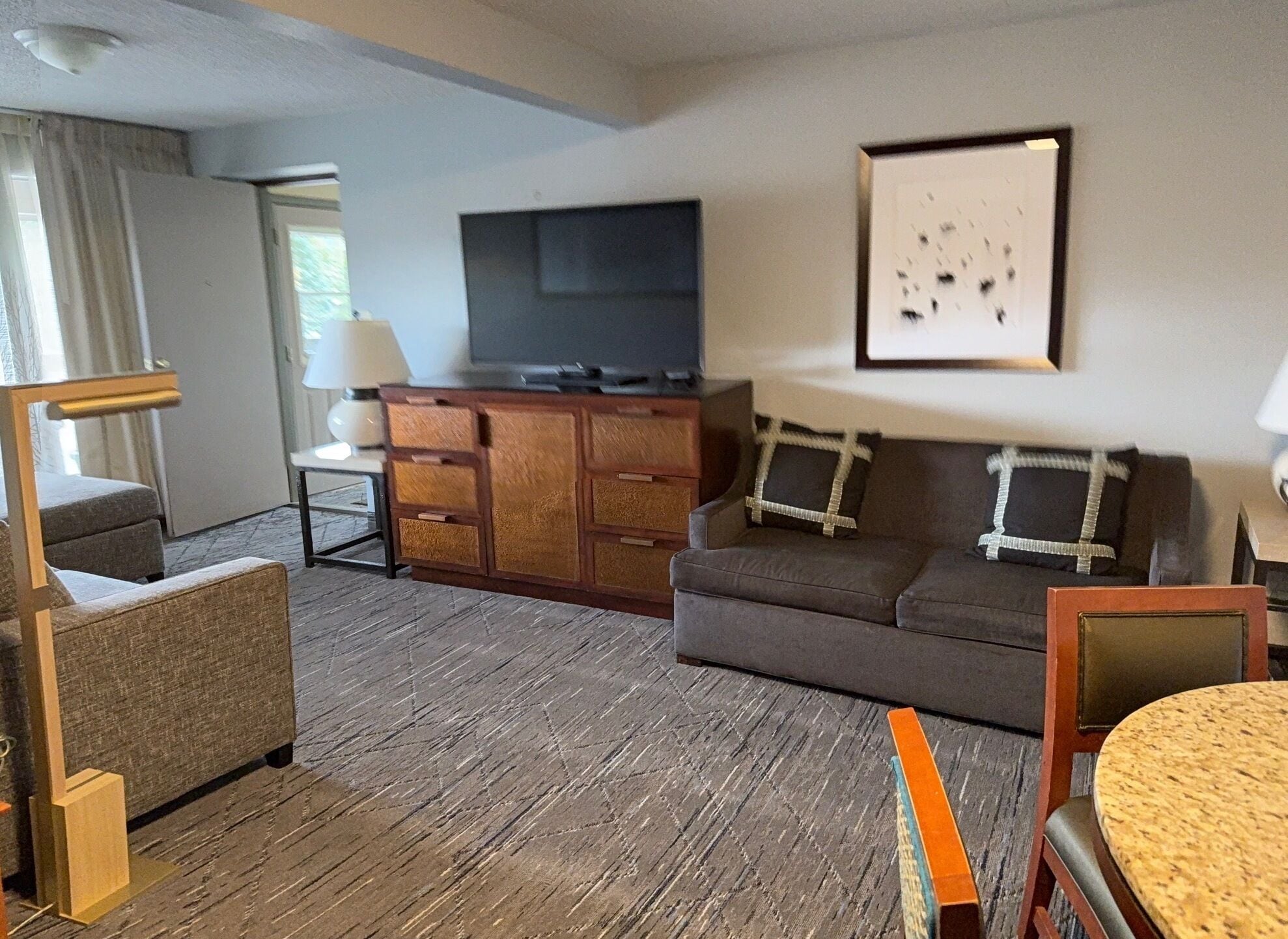 A hotel room with a wooden floor, a sofa, a small table, a TV on a wooden cabinet, and a framed picture on the wall.
