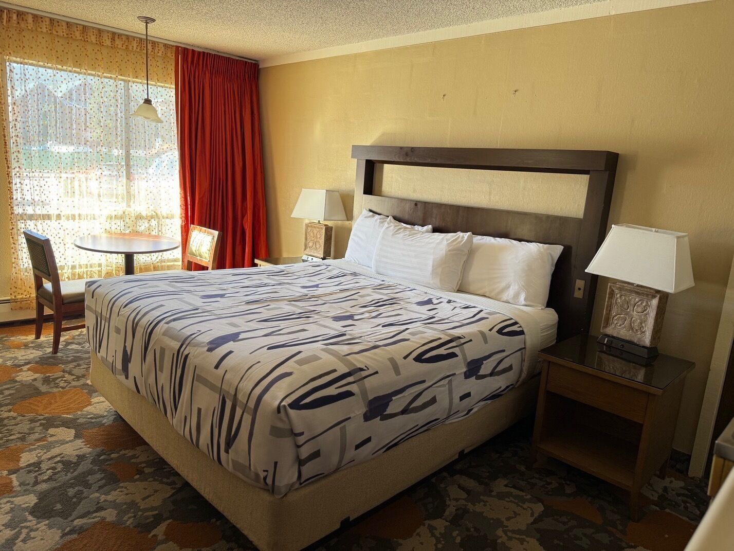 A hotel room with a large bed, two bedside tables, a chair, a small table, and a window with curtains.