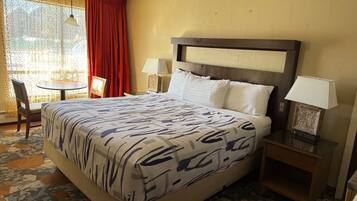A hotel room with a large bed, two bedside tables, a chair, a small table, and a window with curtains.