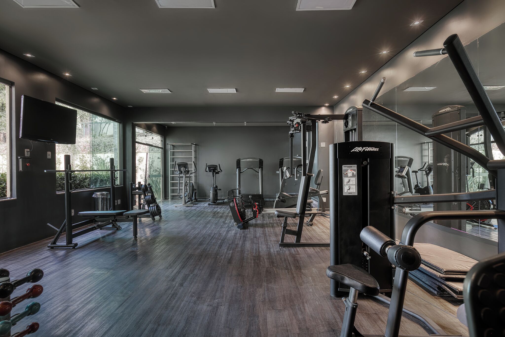 Un gimnasio moderno con diversos equipos de ejercicio, ventanas amplias y un muro con espejos.