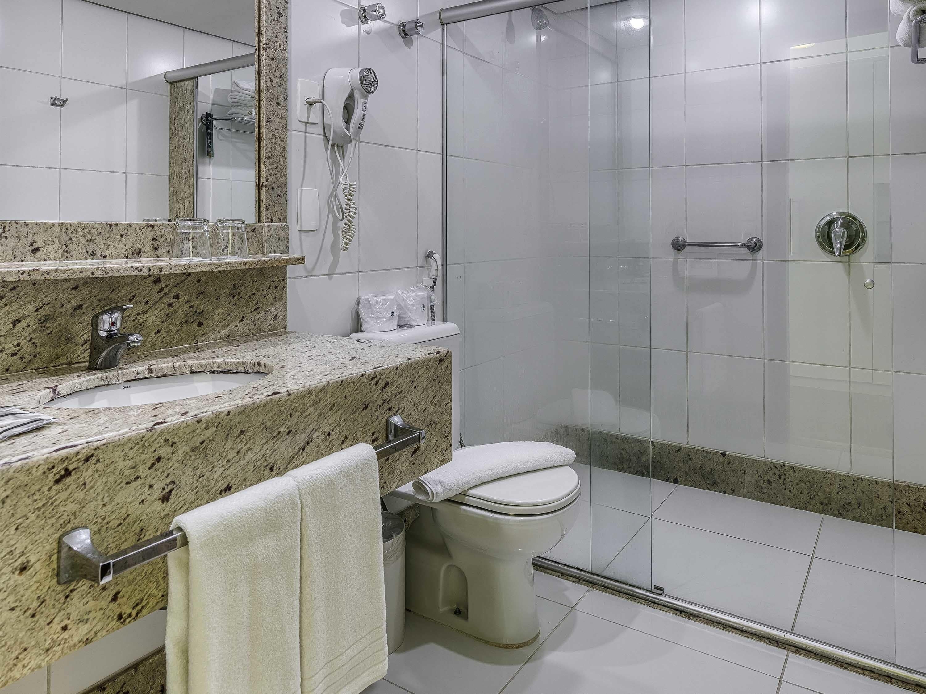 A bathroom with a granite countertop sink, a toilet, a glass shower enclosure, and a hairdryer mounted on the wall.