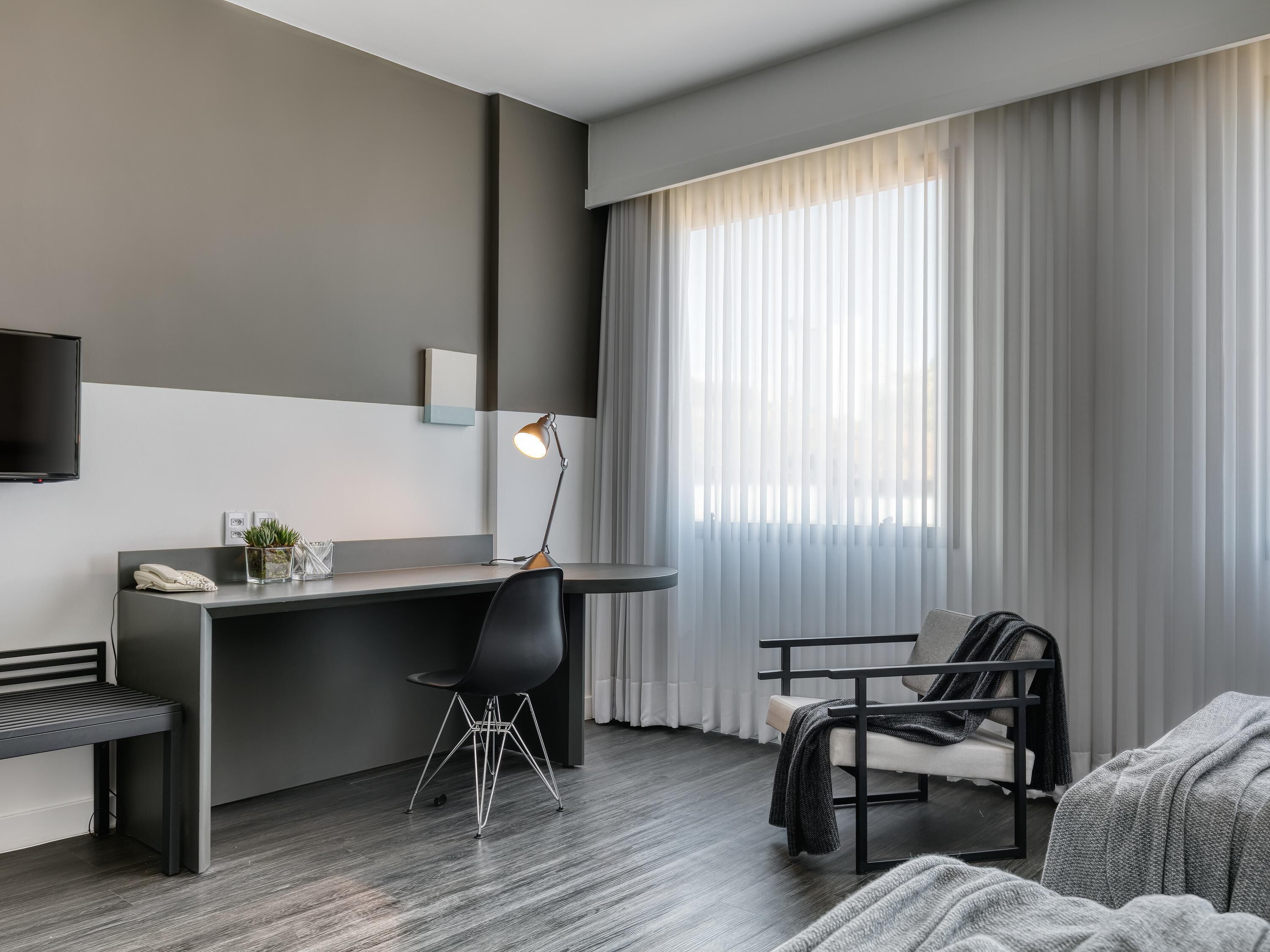 A modern hotel room with a grey and white color scheme, a desk with a lamp, a chair, a sofa, and a large window with sheer curtains.