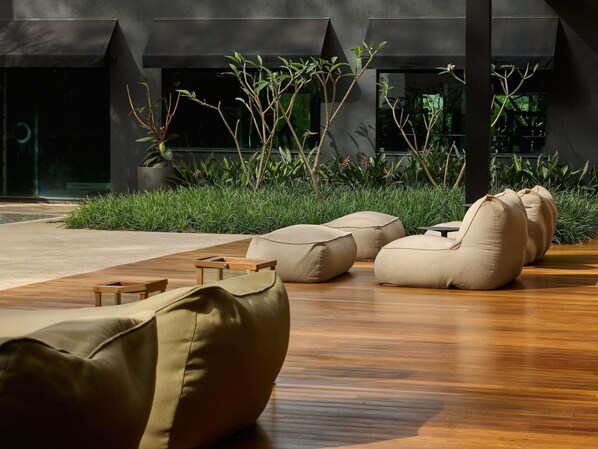 A modern outdoor seating area with bean bags and a wooden table, surrounded by lush greenery and a building with large windows.