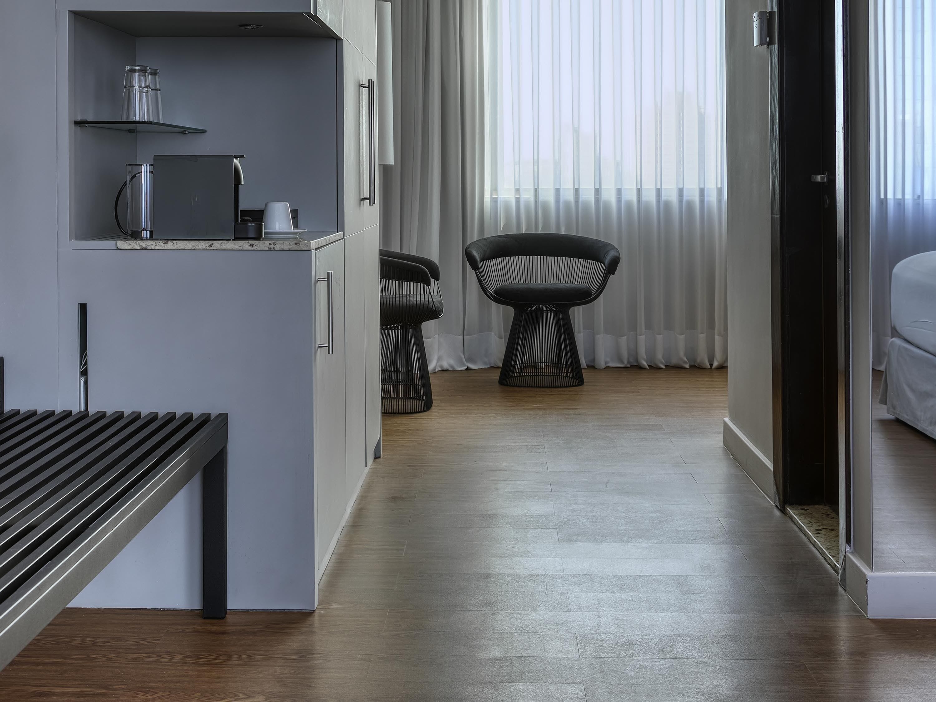 Une chambre d’hôtel moderne avec un plancher en bois, un lit, un bureau avec une chaise, une petite table avec une chaise et une fenêtre avec des rideaux.