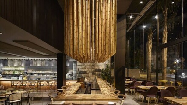 A modern restaurant with a long wooden table and chairs, a hanging bamboo light fixture, and a stone wall in the background. - Caesar Business Belo Horizonte Belvedere Managed by Mercure (Belo Horizonte)