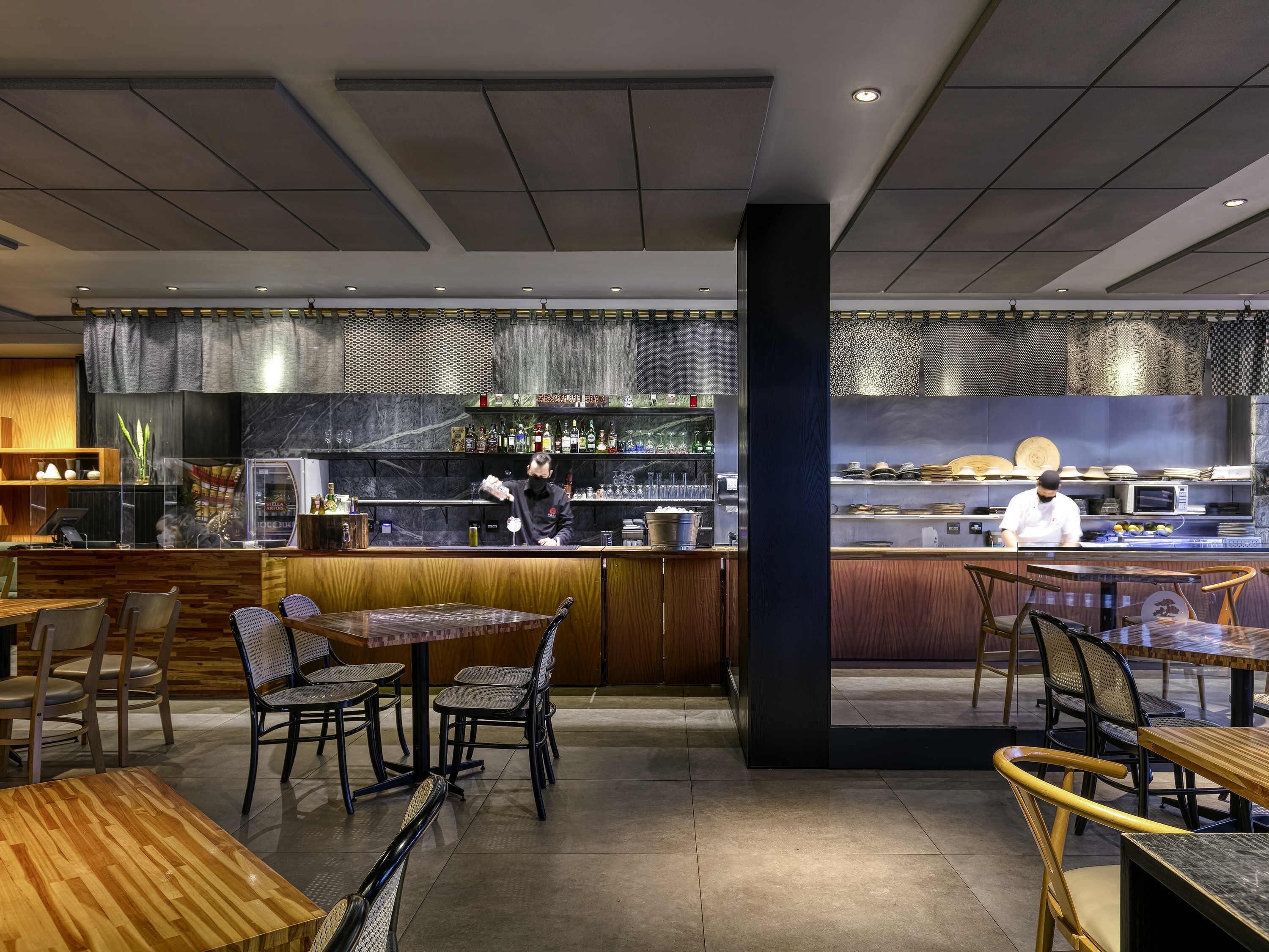 Ein modernes Restaurant mit Holztischen und -stühlen, einer Bar und einer dahinterliegenden Küche.