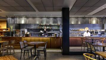 Un interior de restaurante moderno con mesas y sillas de madera, una zona de bar y una cocina al fondo.