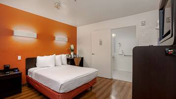 Una habitación de hotel con una cama, una mesita de noche, un escritorio con una silla, una pequeña mesa con un florero y una puerta que da a un baño.