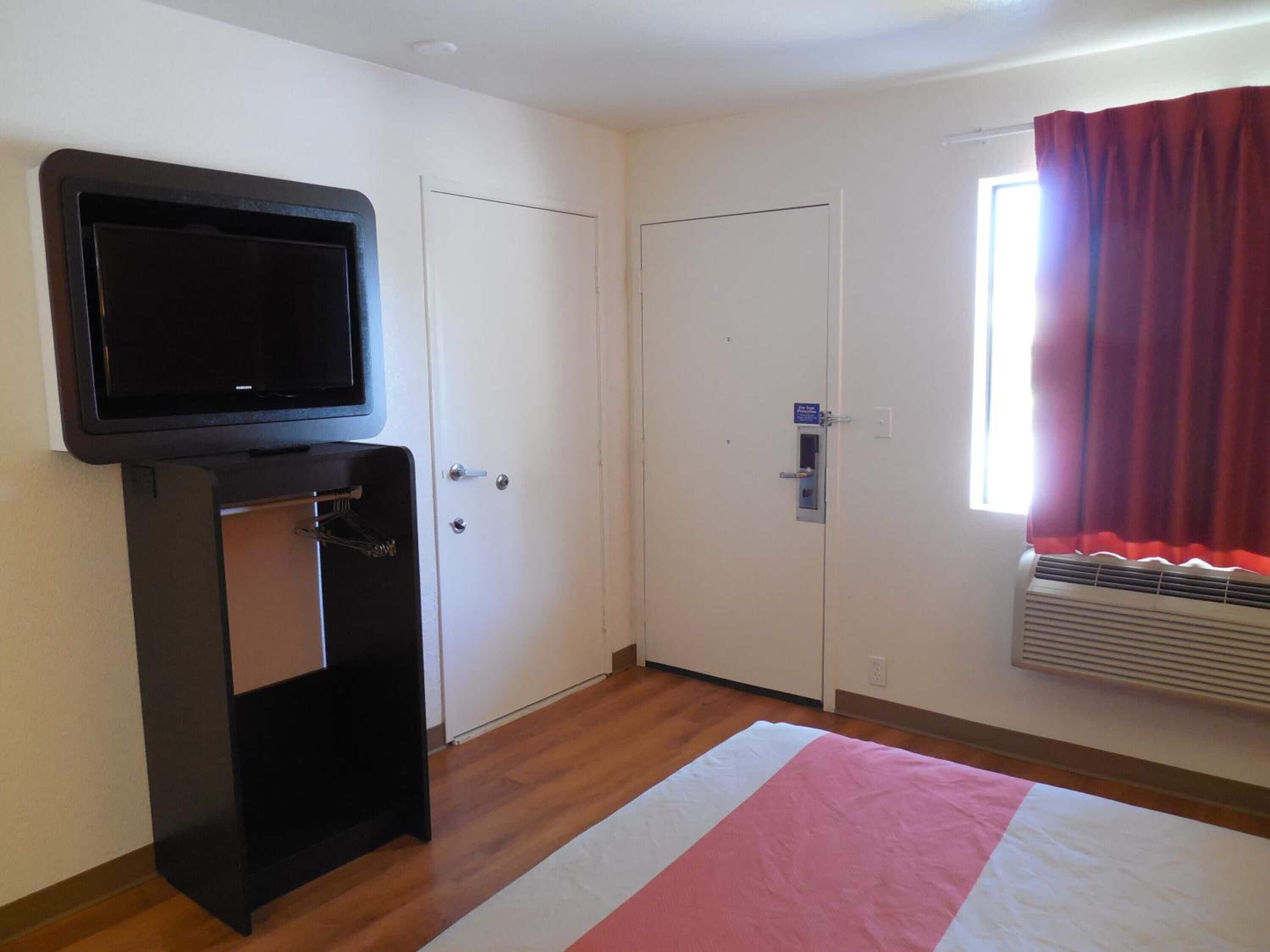 A room with a television mounted on a stand, a door with a security lock, a window with red curtains, and an air conditioning unit.