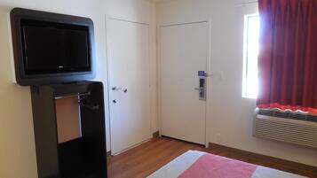 A room with a television mounted on a stand, a door with a security lock, a window with red curtains, and an air conditioning unit.