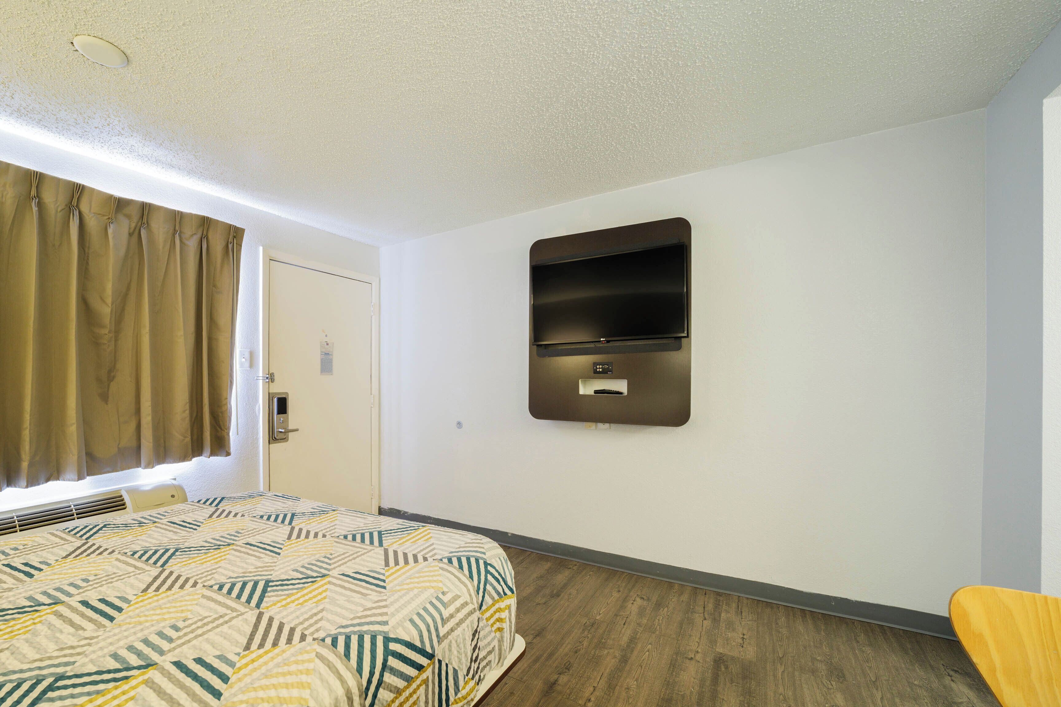 Une chambre d’hôtel avec un lit, un téléviseur fixé au mur, un plancher en bois et une fenêtre avec des rideaux.