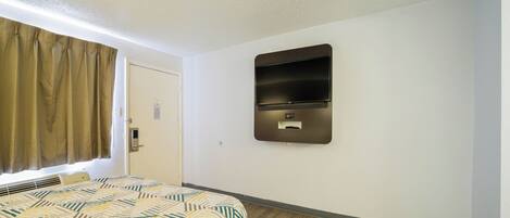 Une chambre d’hôtel avec un lit, un téléviseur fixé au mur, un plancher en bois et une fenêtre avec des rideaux.