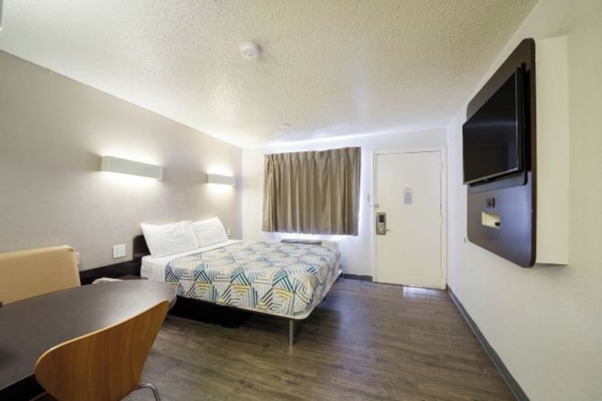 A hotel room with a bed, a desk, a chair, a TV mounted on the wall, and a door.