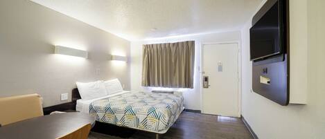 Quarto de hotel com cama, escrivaninha, cadeira, televisão e janela com cortinas.
