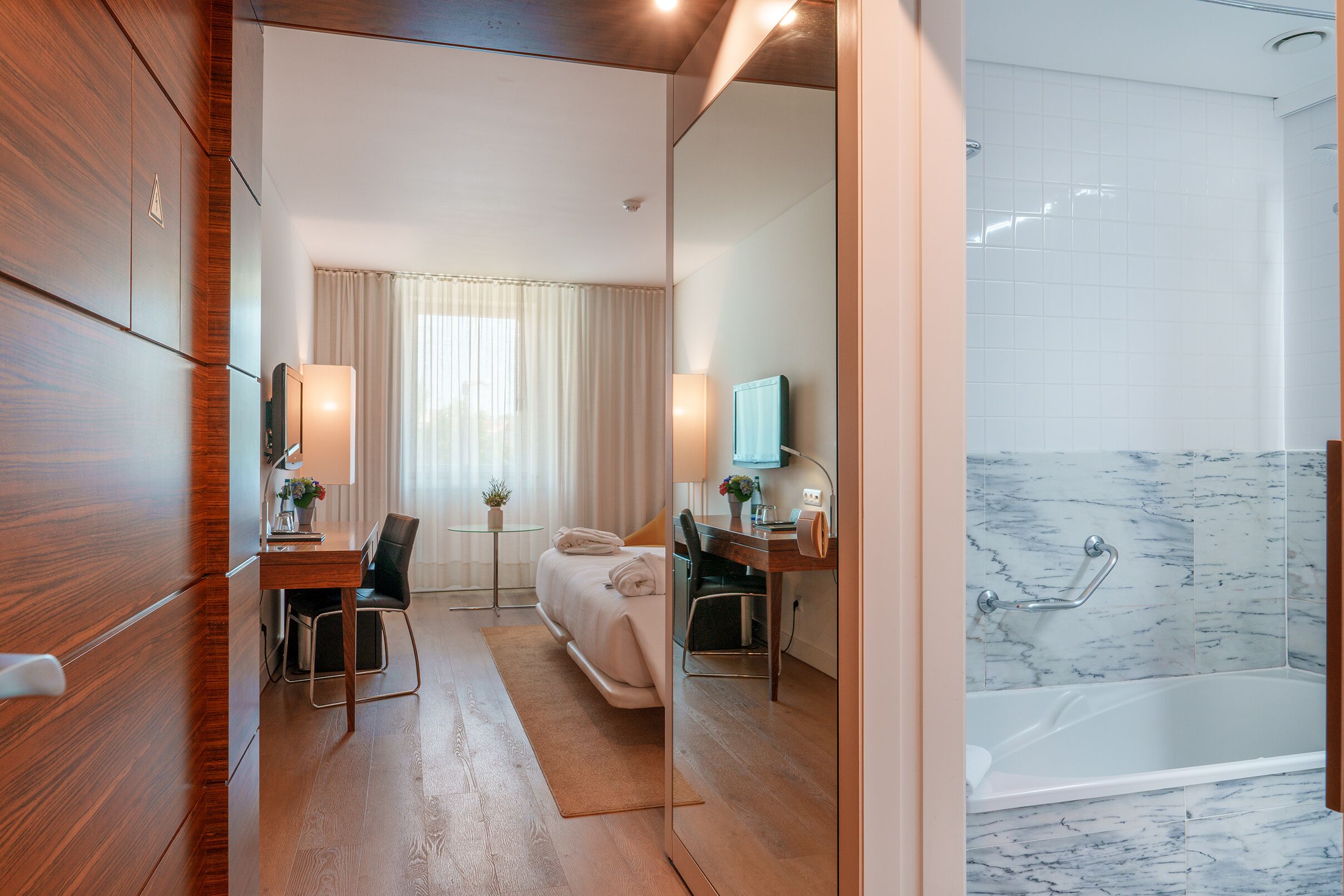 A hotel room with a wooden floor, a bed, a desk with a chair, a bathroom with a bathtub, and a large mirror.