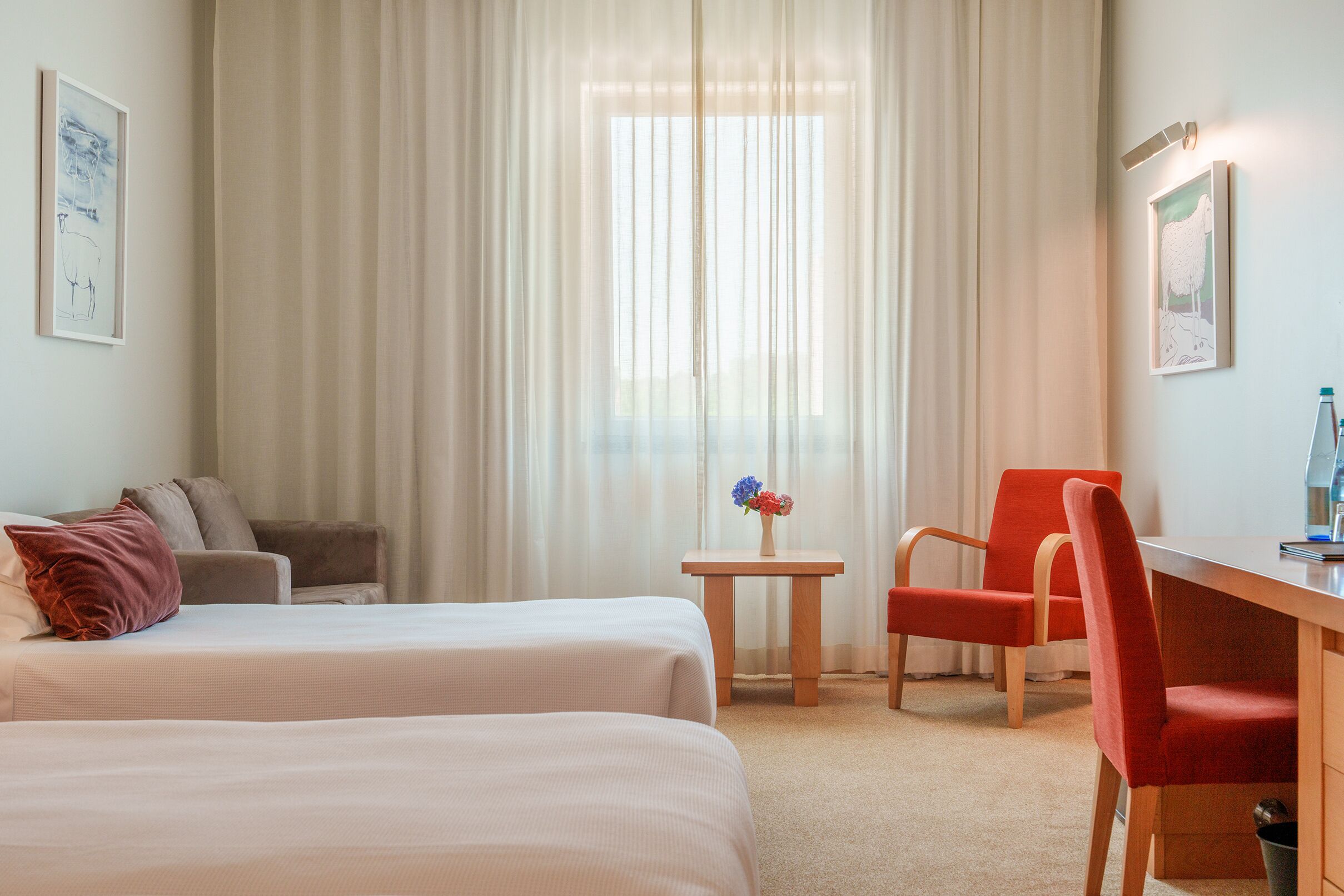 A hotel room with two beds, a desk, a chair, a small table with a vase, and a painting on the wall.