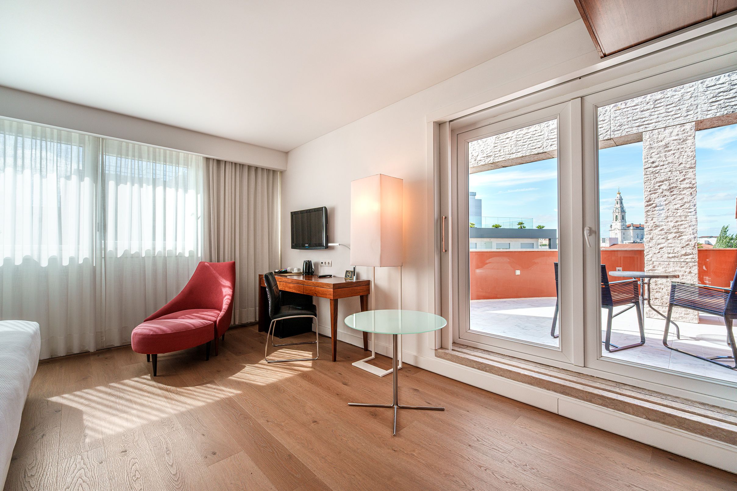 Habitación de hotel con una silla roja, una mesita pequeña, un televisor en la pared y vistas de la ciudad a través de un ventanal.